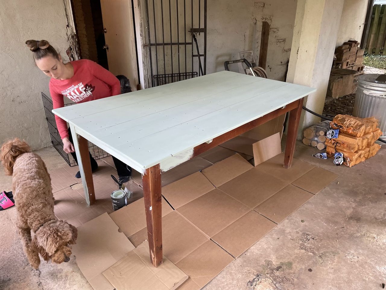 A woman with light-brown hair painting a wooden dining table with pale green chalk paint outdoors, while Remi looks on nearby