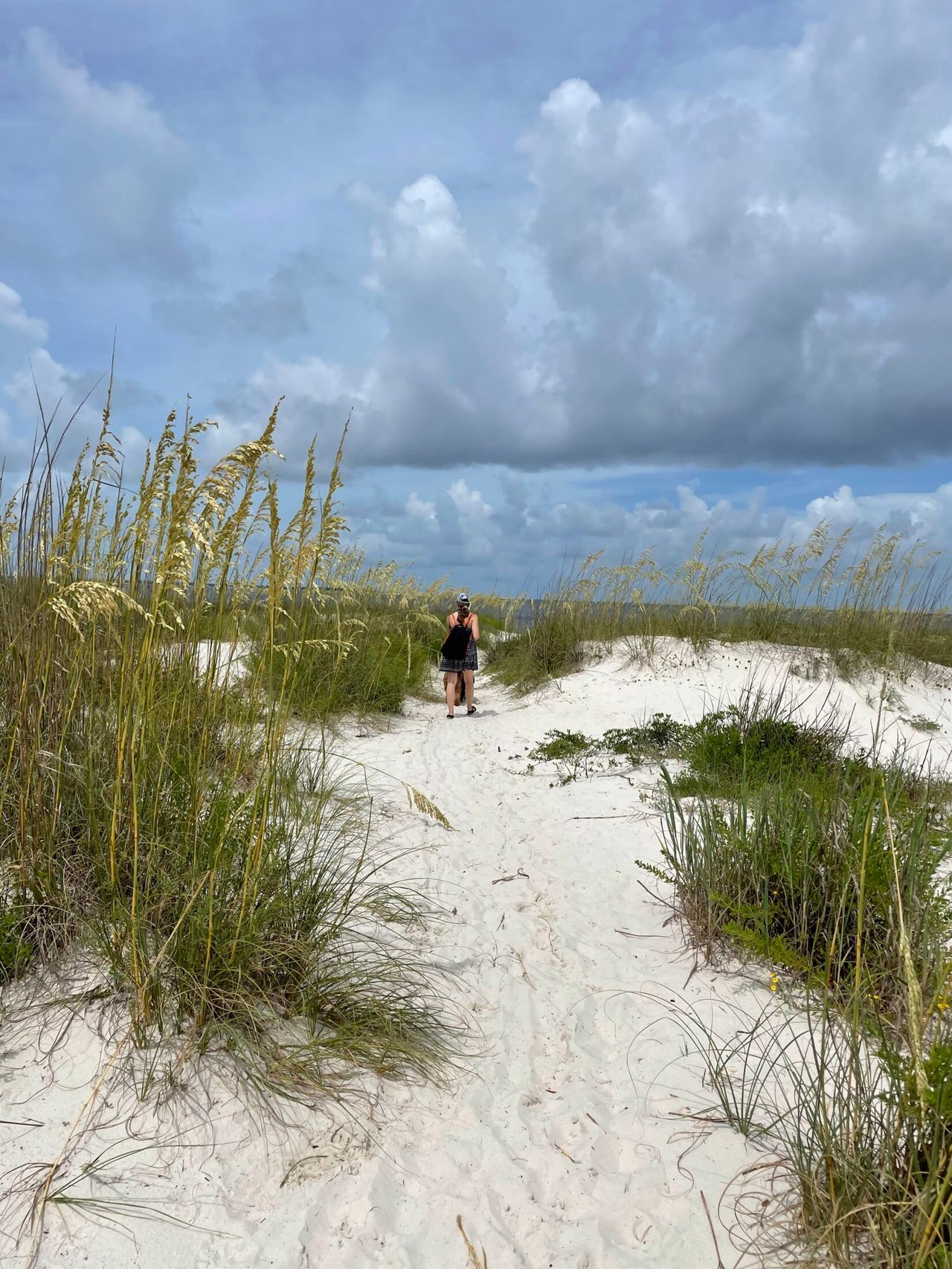 A woman walks away along a white sand path through sea oats and dune grass under a cloudy blue sky