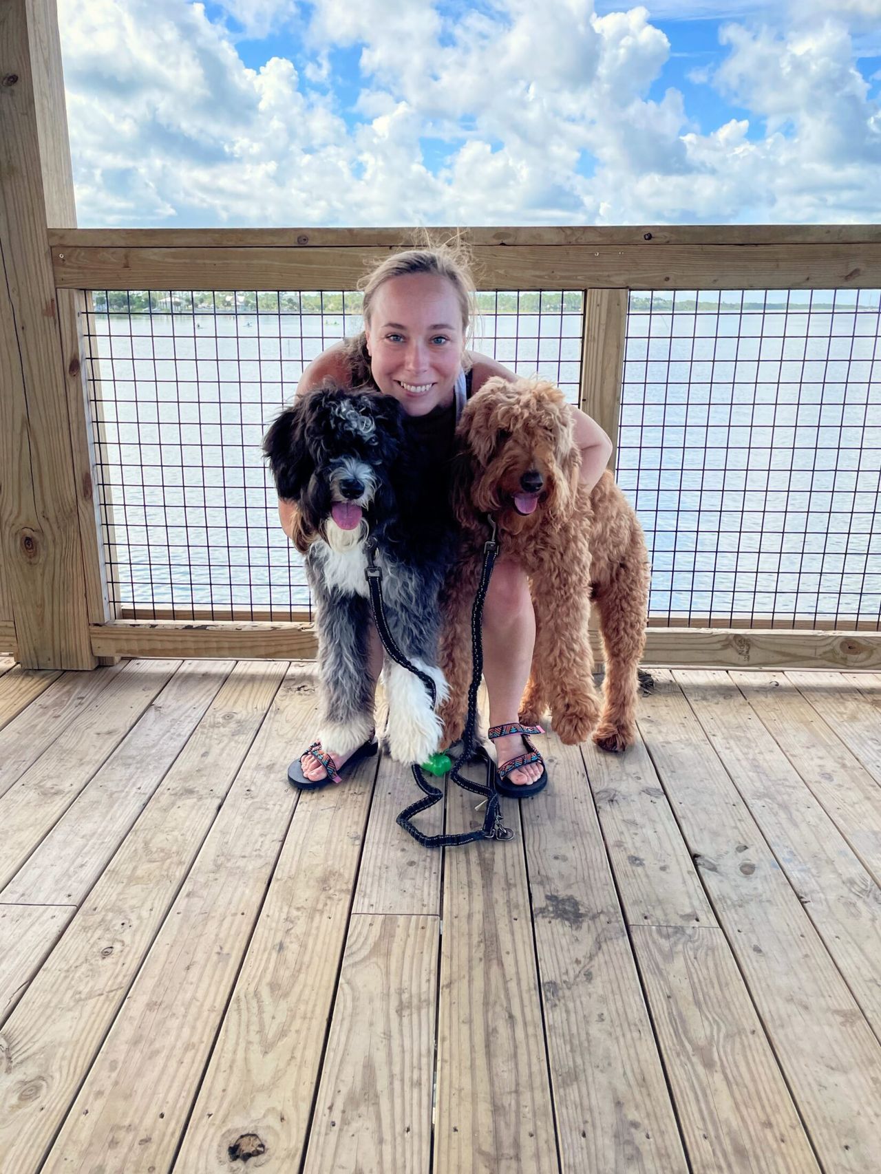 A woman with light-brown hair kneels on a waterfront deck hugging Otto and Remi, with blue sky and water in the background