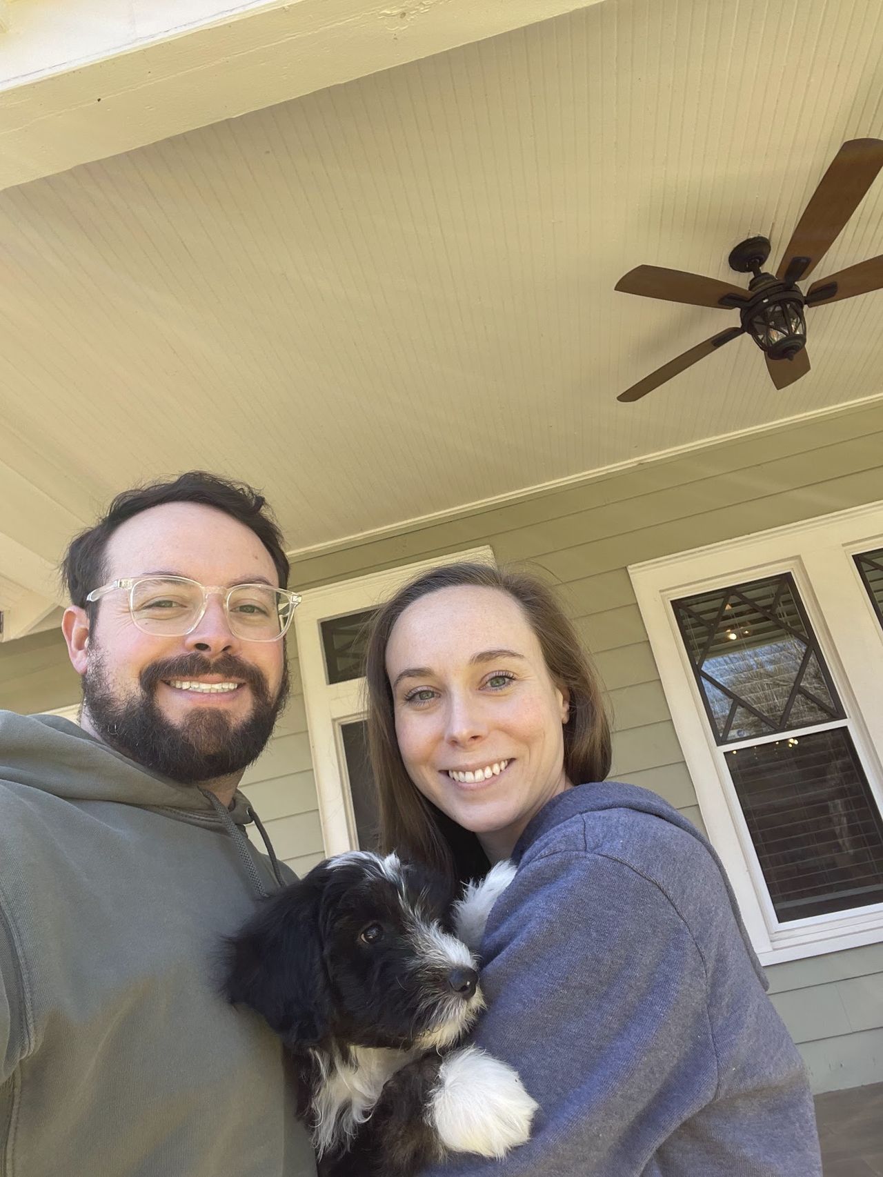 Sam Solomon and a woman with light-brown hair smile on a covered porch while holding a small black and white puppy