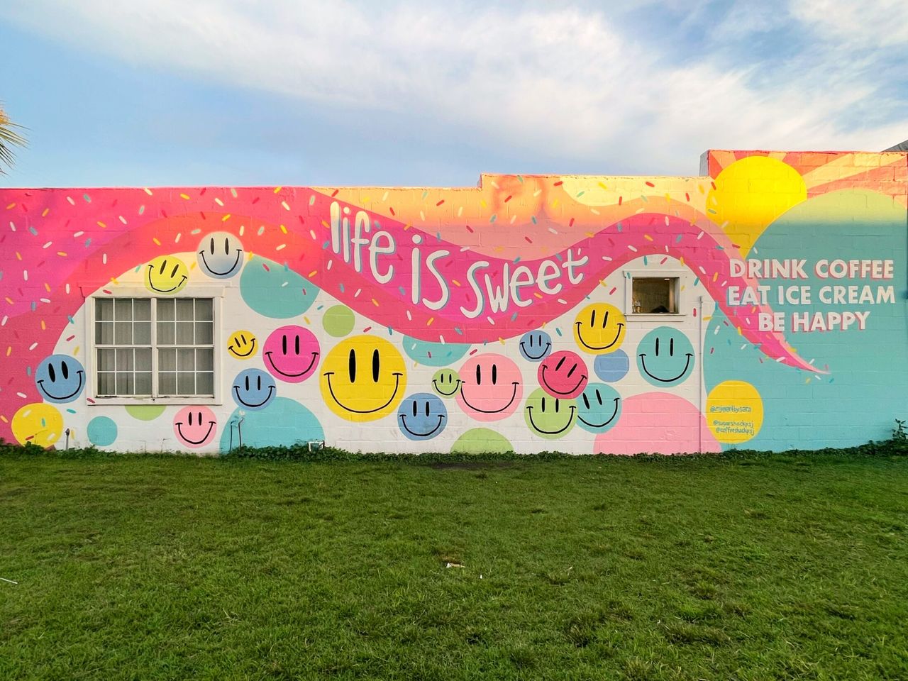 Colorful outdoor mural reading 'life is sweet / Drink Coffee Eat Ice Cream Be Happy' with smiley faces, confetti, and pastel circles on a building exterior