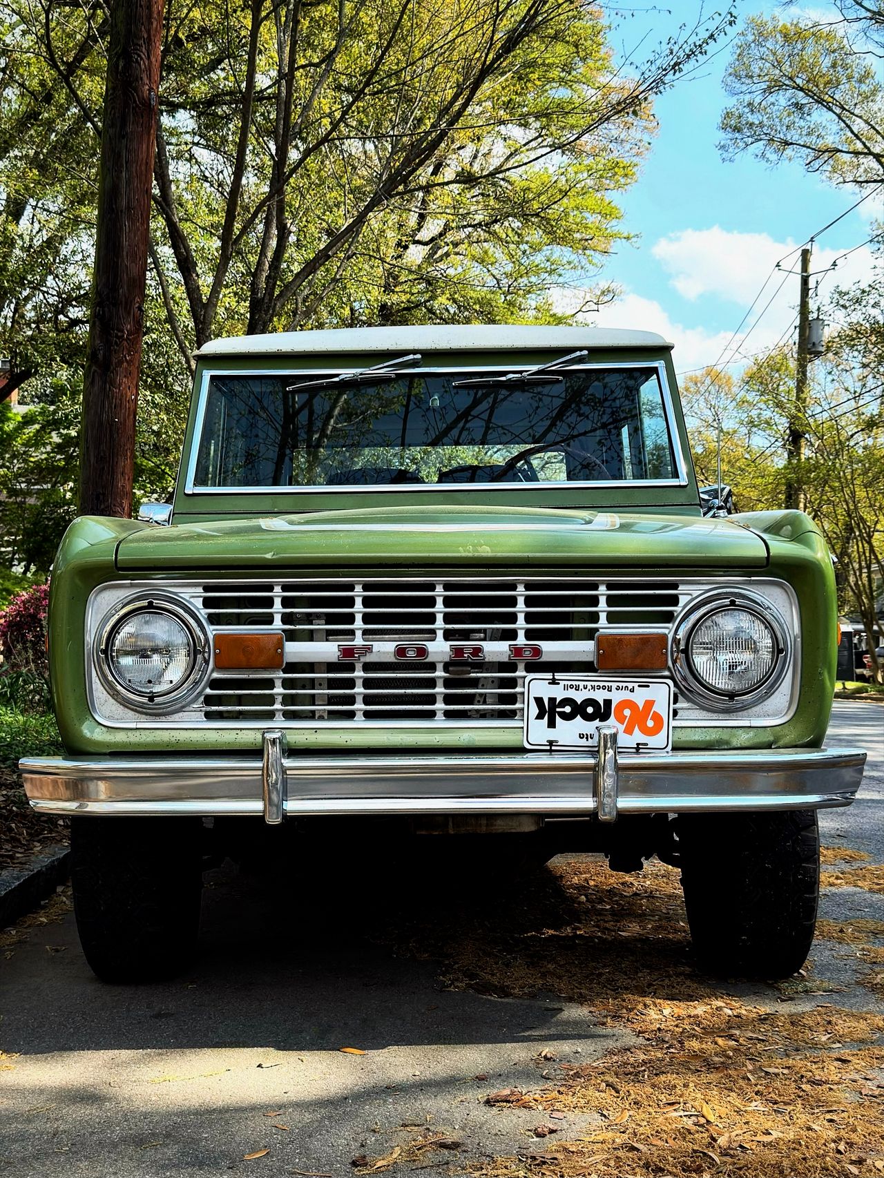 Straight-on front view of a green vintage Ford Bronco with chrome grille and FORD lettering, parked under trees.