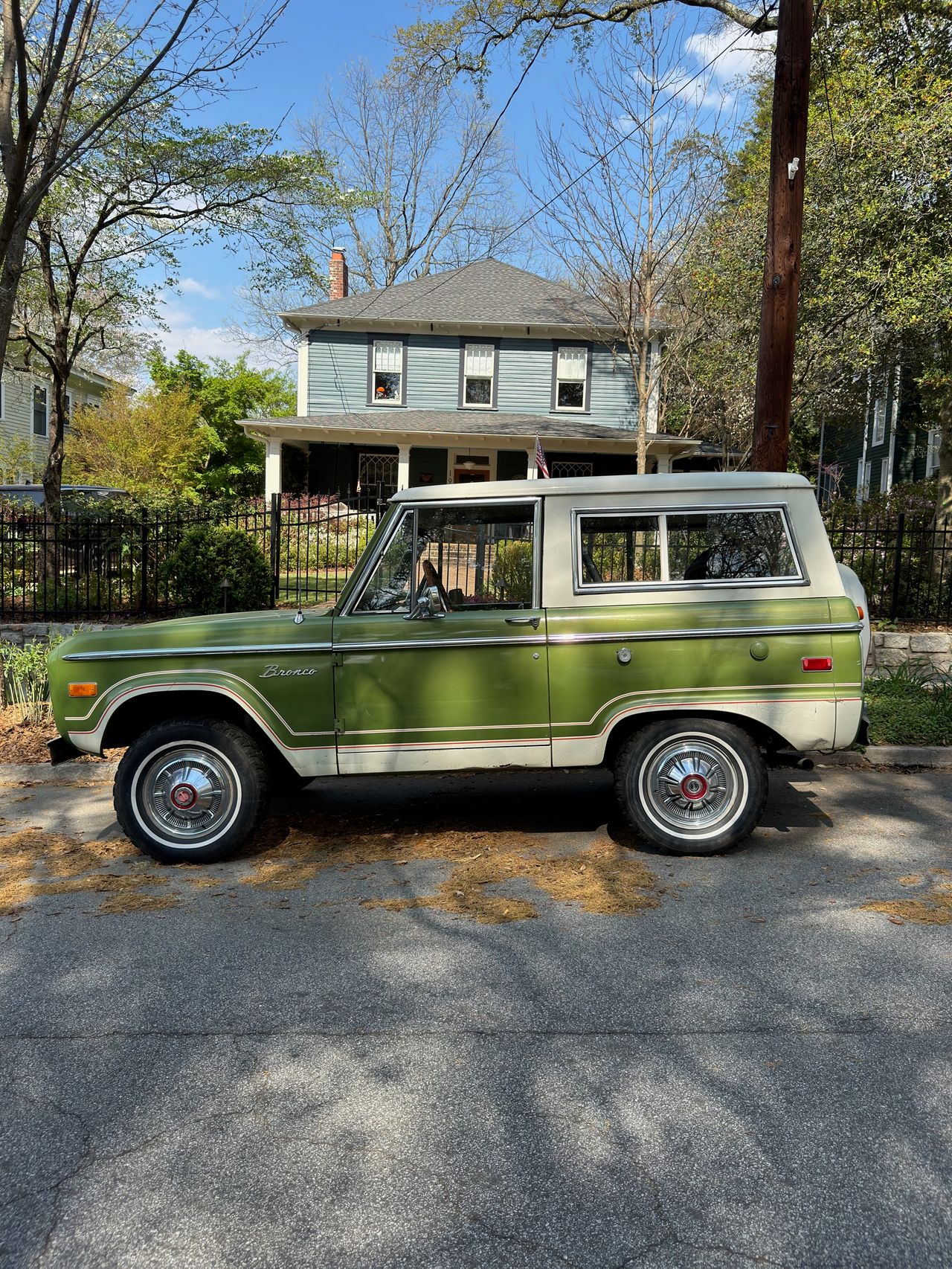 Side profile of a green and cream vintage Ford Bronco parked along a residential street with a Victorian house behind.