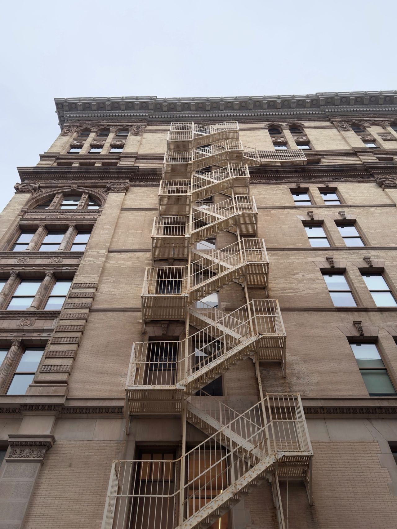 A cream-painted fire escape zigzags up the front of a tan brick SoHo building with ornate window trim.
