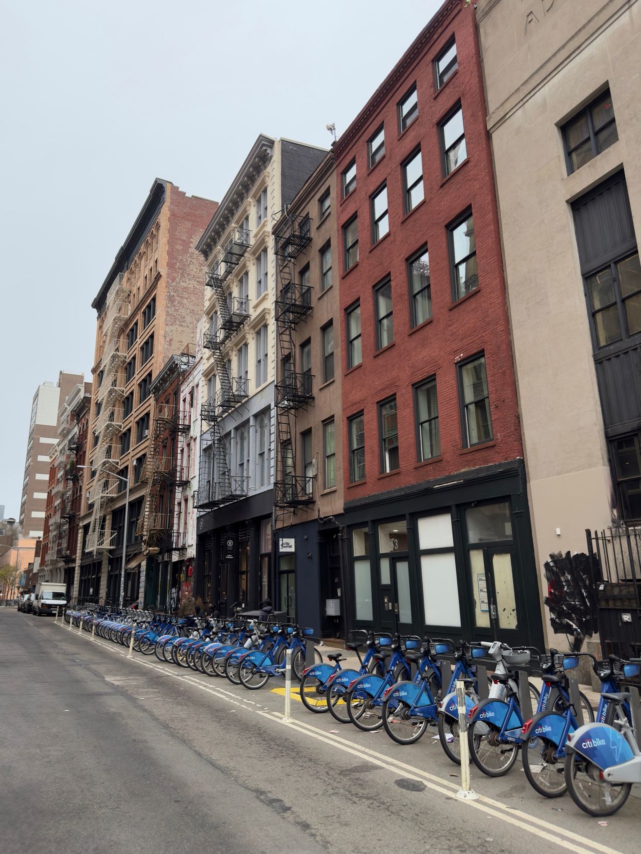 A long row of blue Citi Bikes parked along a SoHo street lined with brick buildings and black fire escapes.