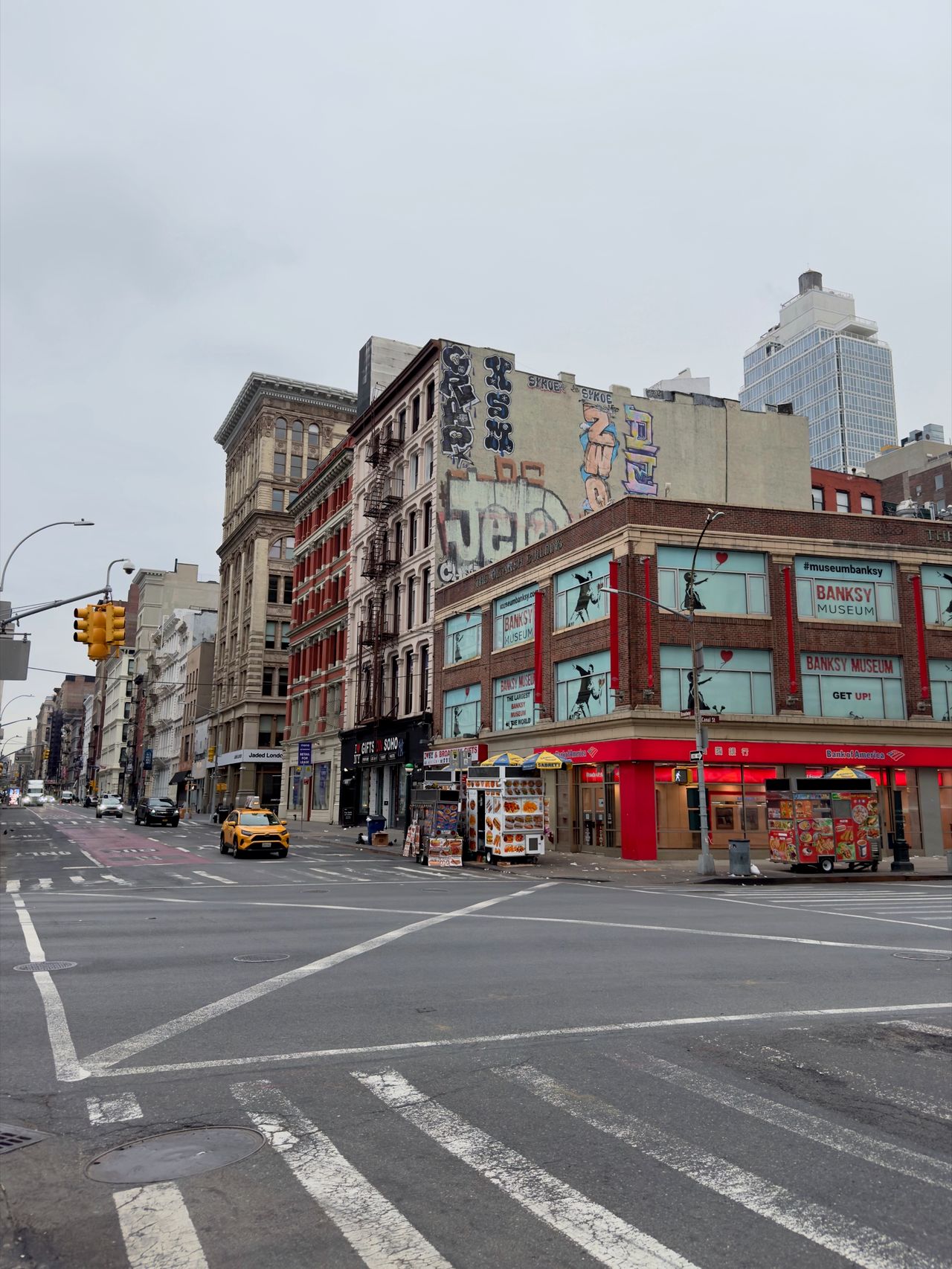 A SoHo street corner with a yellow taxi, food carts, and a building advertising the Banksy Museum.