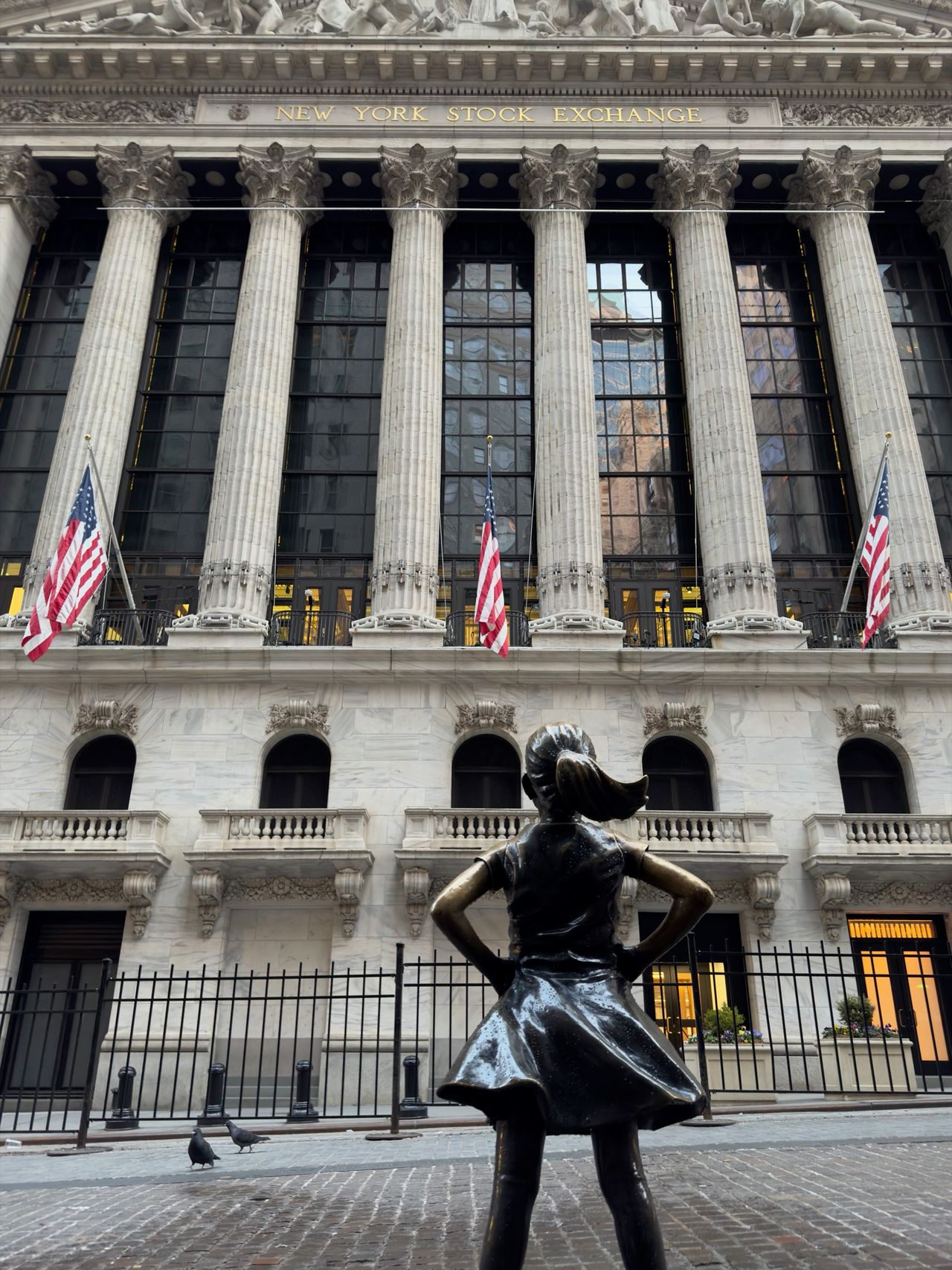 A small bronze statue of a girl in a dress stands with her hands on her hips, facing the columned facade of the New York Stock Exchange and its three American flags.