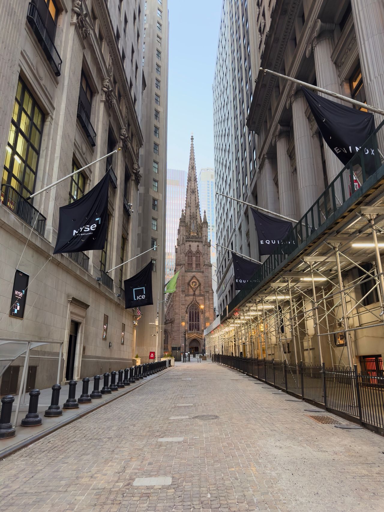 Looking down Wall Street between tall buildings toward the spire of Trinity Church, with NYSE banners on either side.