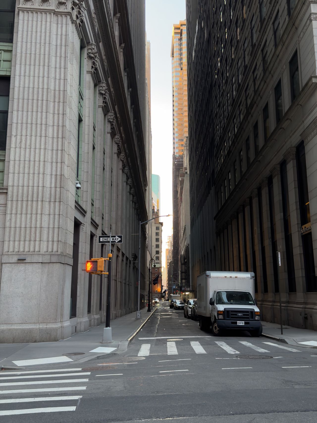 A narrow canyon between Wall Street buildings with classical columns on the left, looking toward distant skyscrapers.