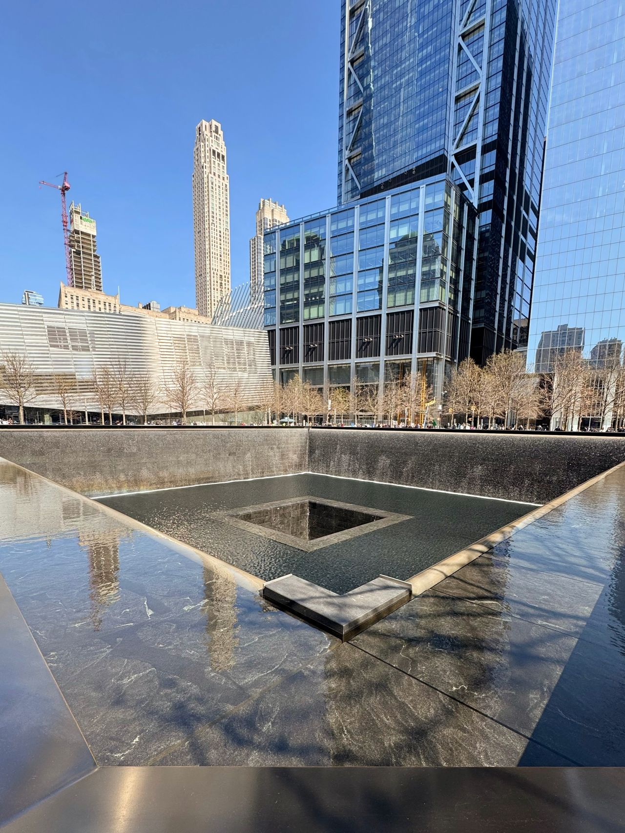 A square reflecting pool at the 9/11 Memorial, water cascading inward, with One World Trade Center and neighboring towers behind.
