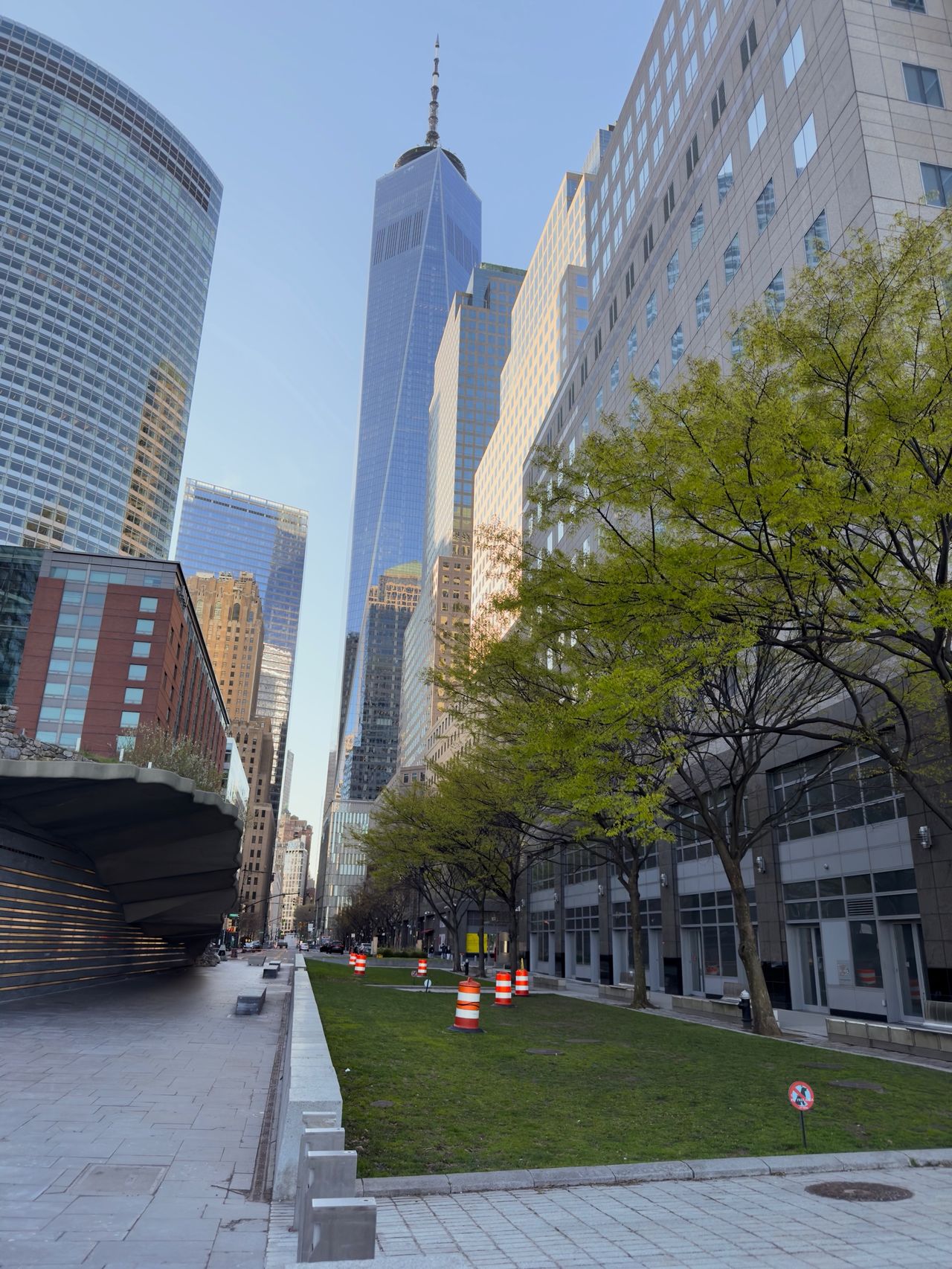 One World Trade Center rises beyond a tree-lined median between two modern streets in lower Manhattan.