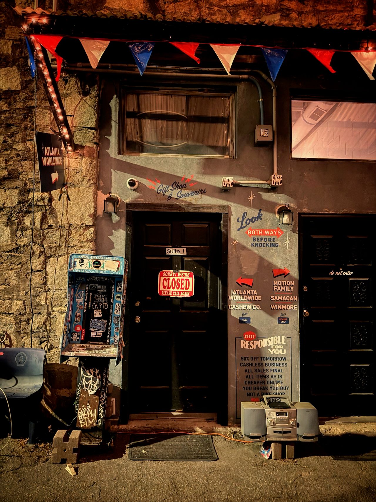 A weathered storefront door at night with a red 'Sorry We're Closed' sign, red and blue triangle bunting hanging above, surrounded by hand-painted shop signs and a graffitied newsbox outside.