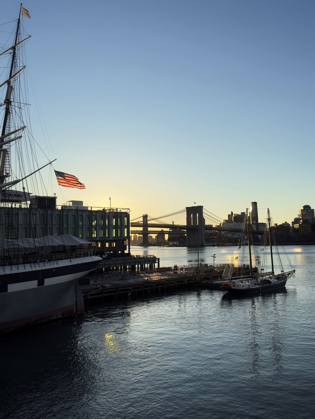 An American flag flies from a tall ship docked at South Street Seaport, with the Brooklyn Bridge silhouetted across the East River and a sailboat moored nearby.