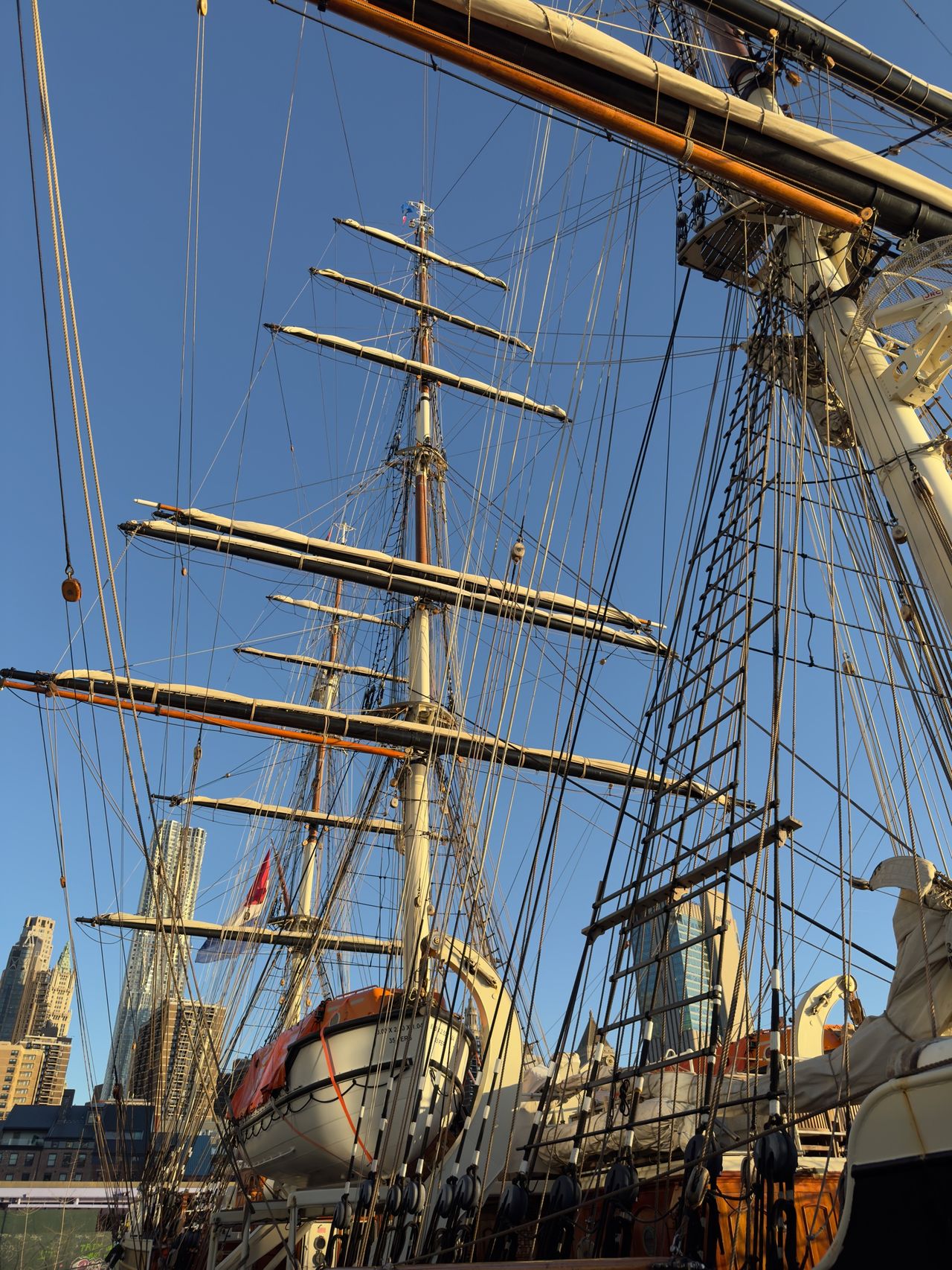 The masts, yards, and rigging of a tall ship rise into a clear blue sky above the lower Manhattan skyline.