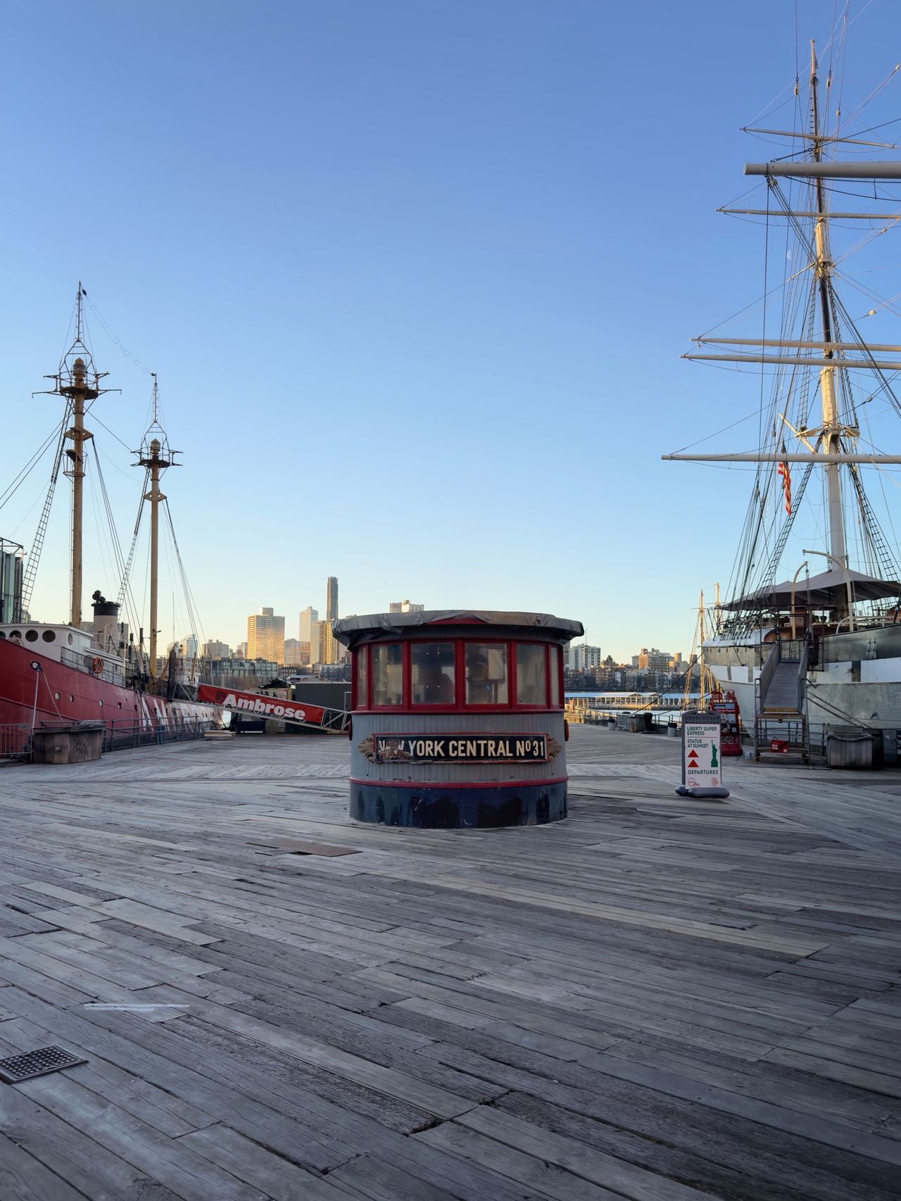 The wooden pier deck of South Street Seaport with the rounded cabin of the York Central No. 31 tugboat, flanked by a red lightship on the left and a tall ship on the right.