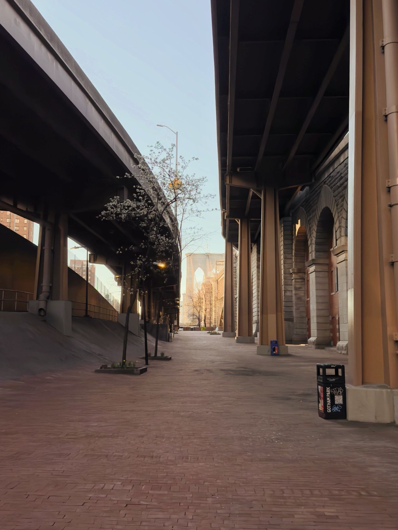 A pedestrian plaza runs between the underside of an elevated highway and a stone-arched colonnade, with a Brooklyn Bridge tower at the far end.