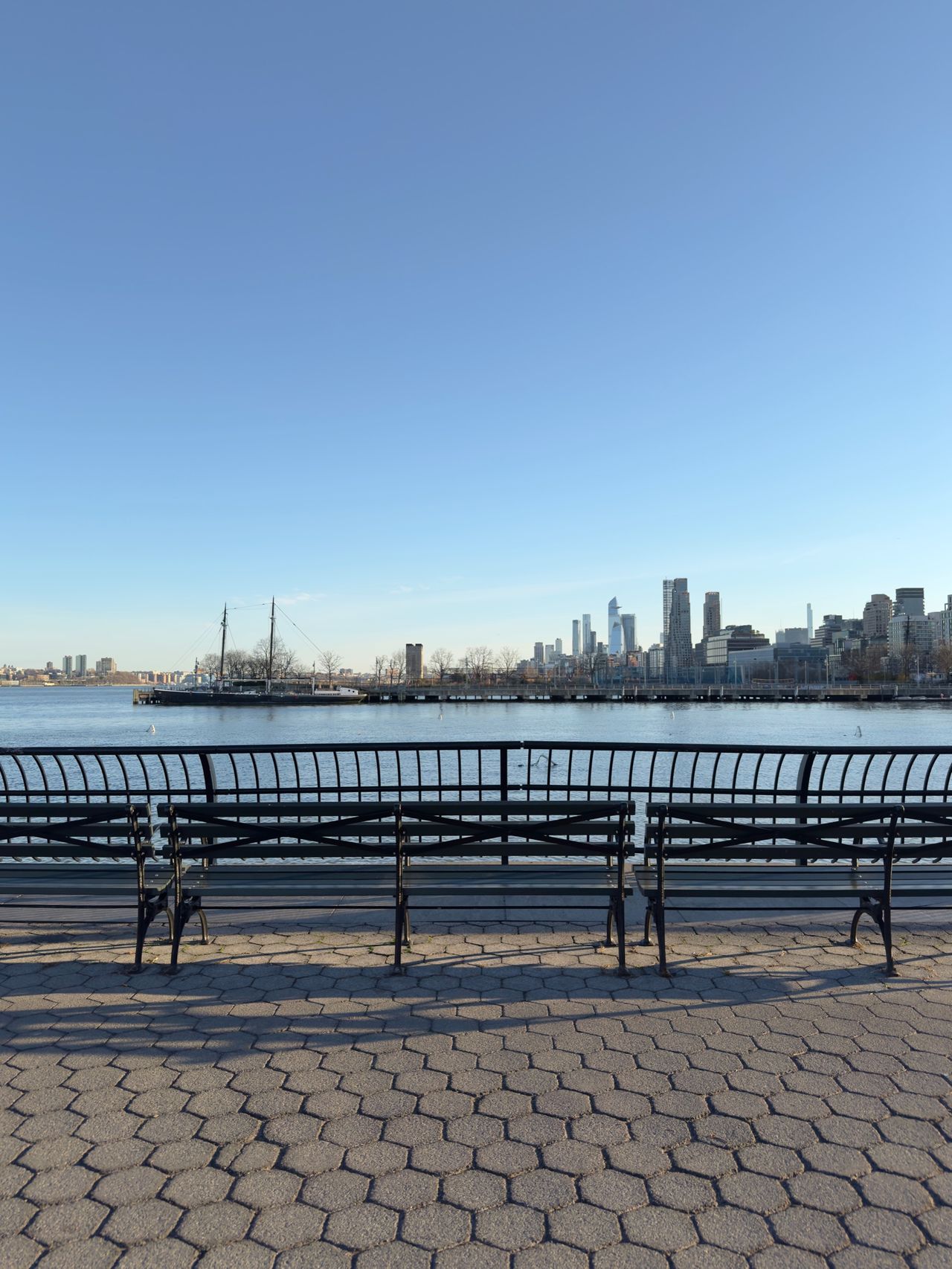 Empty bench on a Hudson River esplanade facing a tall ship moored at a pier with the lower Manhattan skyline beyond