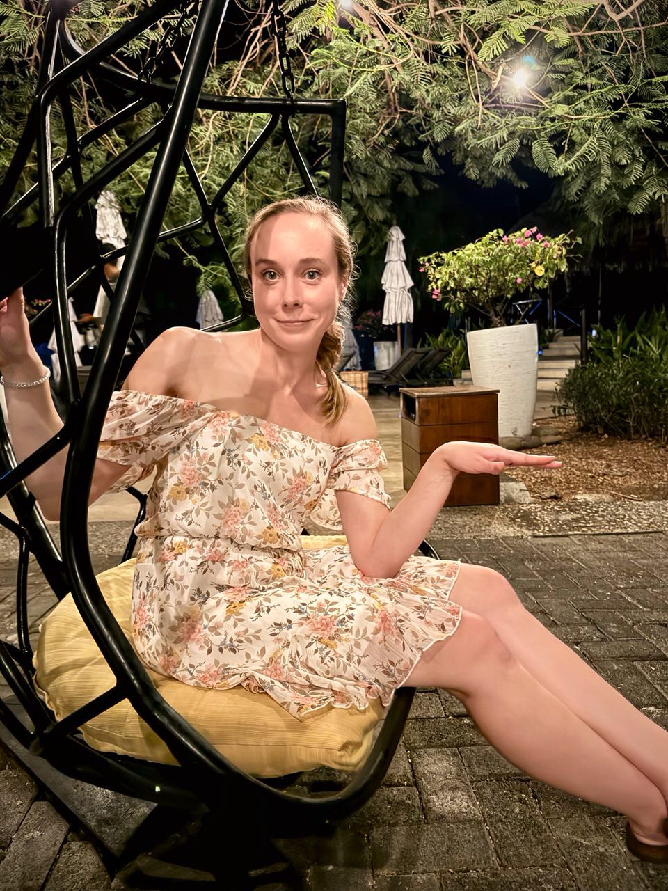 A woman with light-brown hair in a floral off-shoulder dress sits in a hanging egg chair at an outdoor patio at night