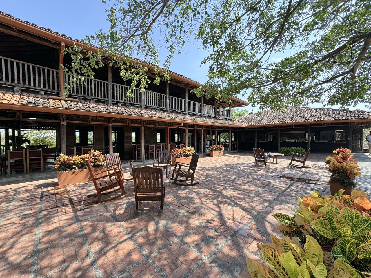 Open-air courtyard with brick pavers, wooden rocking chairs, colorful potted plants, and a two-story hacienda-style building with terracotta roof tiles
