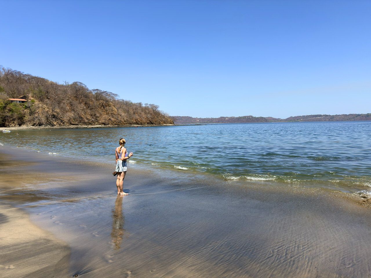 A woman with light-brown hair stands barefoot at the water's edge on a sandy beach, looking out at a calm bay with tree-covered hills beyond