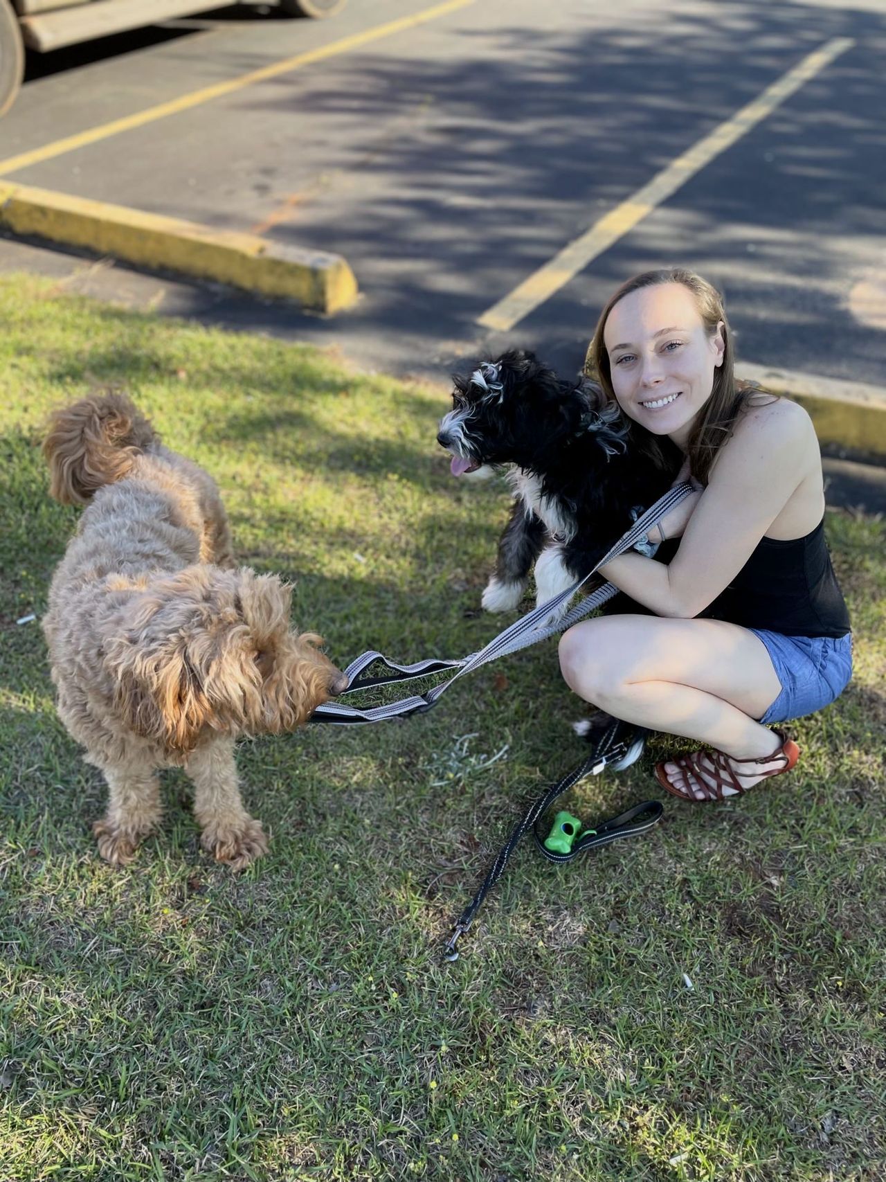 Holly crouches in a black top, holding Otto, a sheepadoodle puppy tightly. Otto has his tongue out. Remi looks at both of them skeptically.