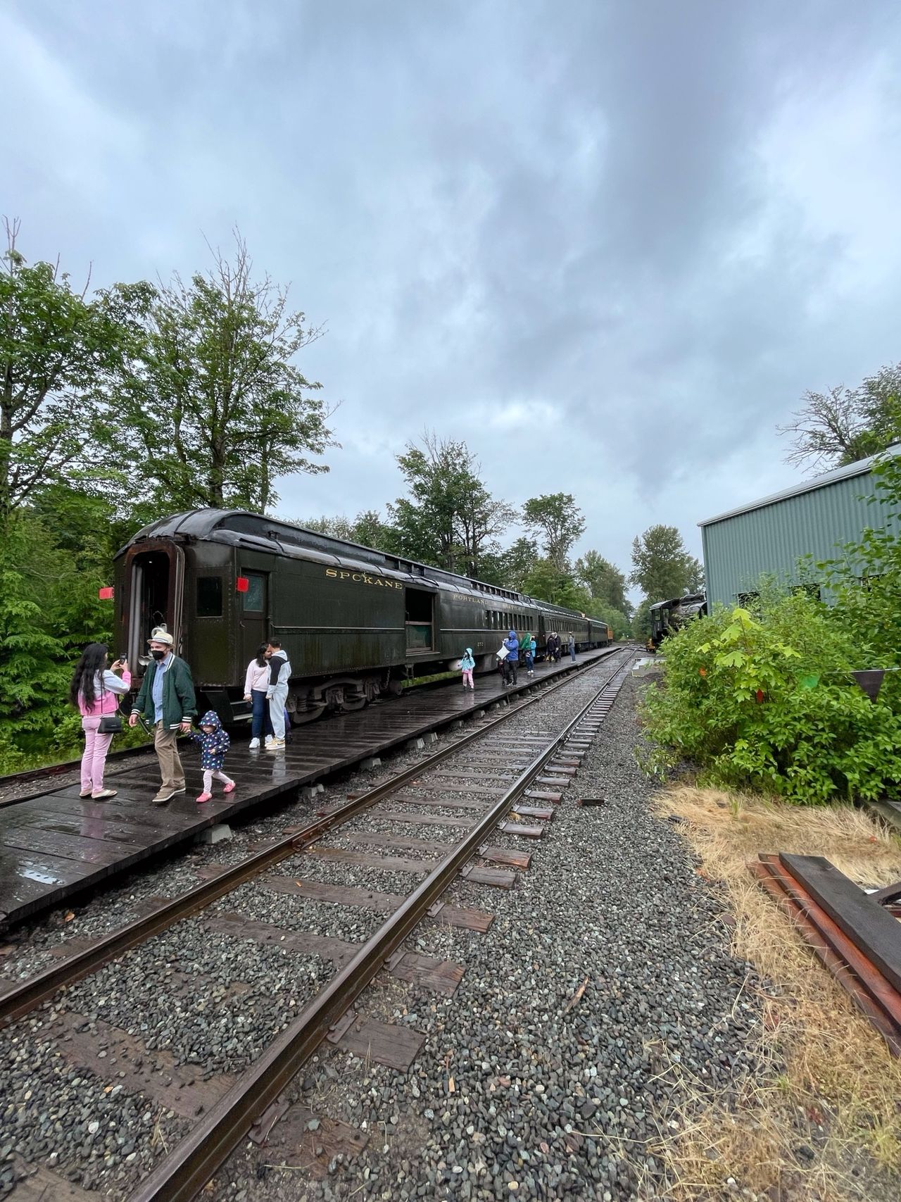 Passengers disembark from vintage Spokane railway cars on a cloudy day. The train, a relic of a bygone era, sits on the tracks surrounded by lush greenery. A group of visitors explore the area with curiosity, some walking along the wooden platform beside the train. The overcast sky creates a dramatic backdrop to this scene of travel and exploration, evoking a sense of nostalgia and adventure.