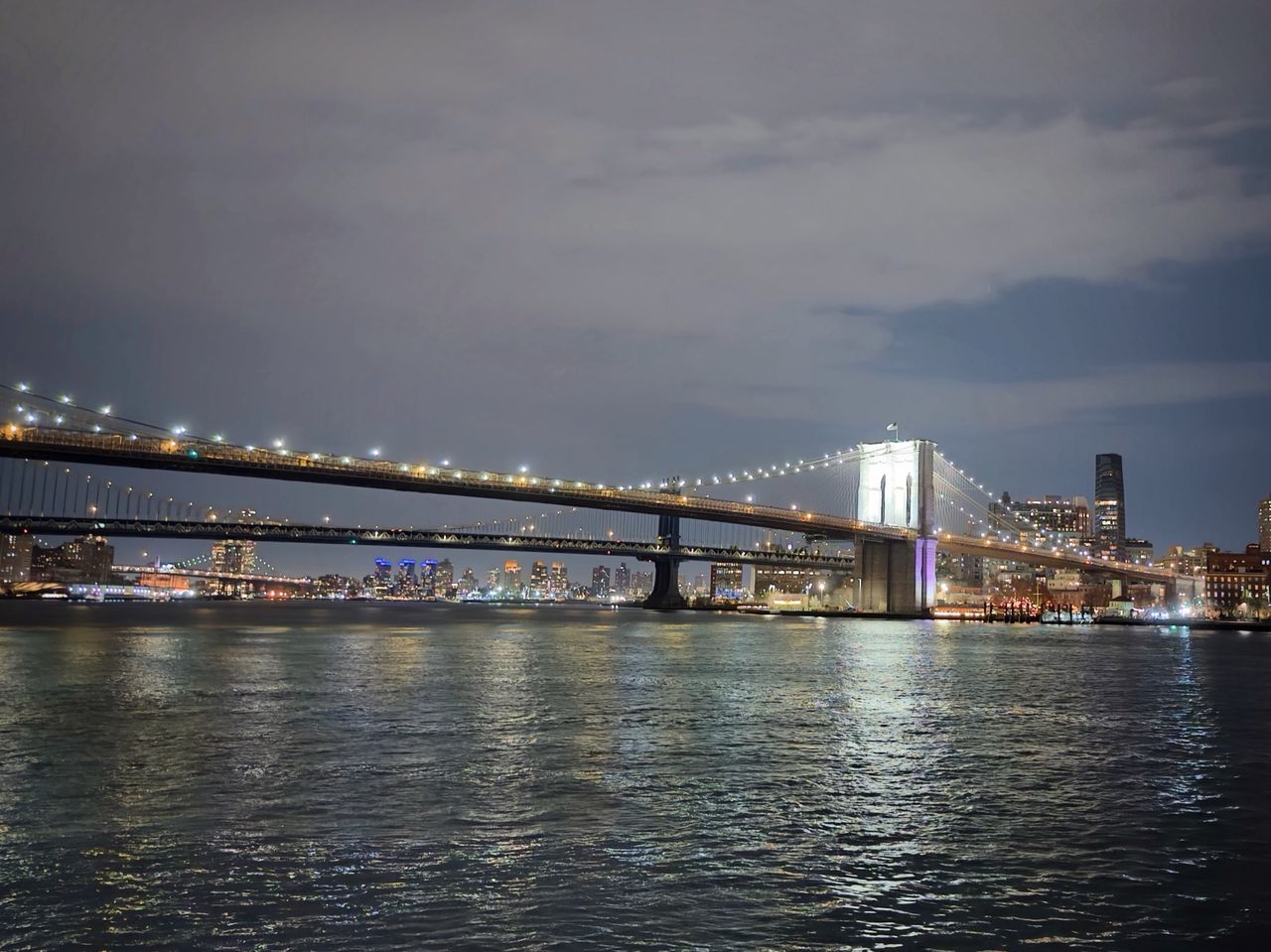 Brooklyn and Manhattan bridges lit up at night across the East River, lights reflecting on the water