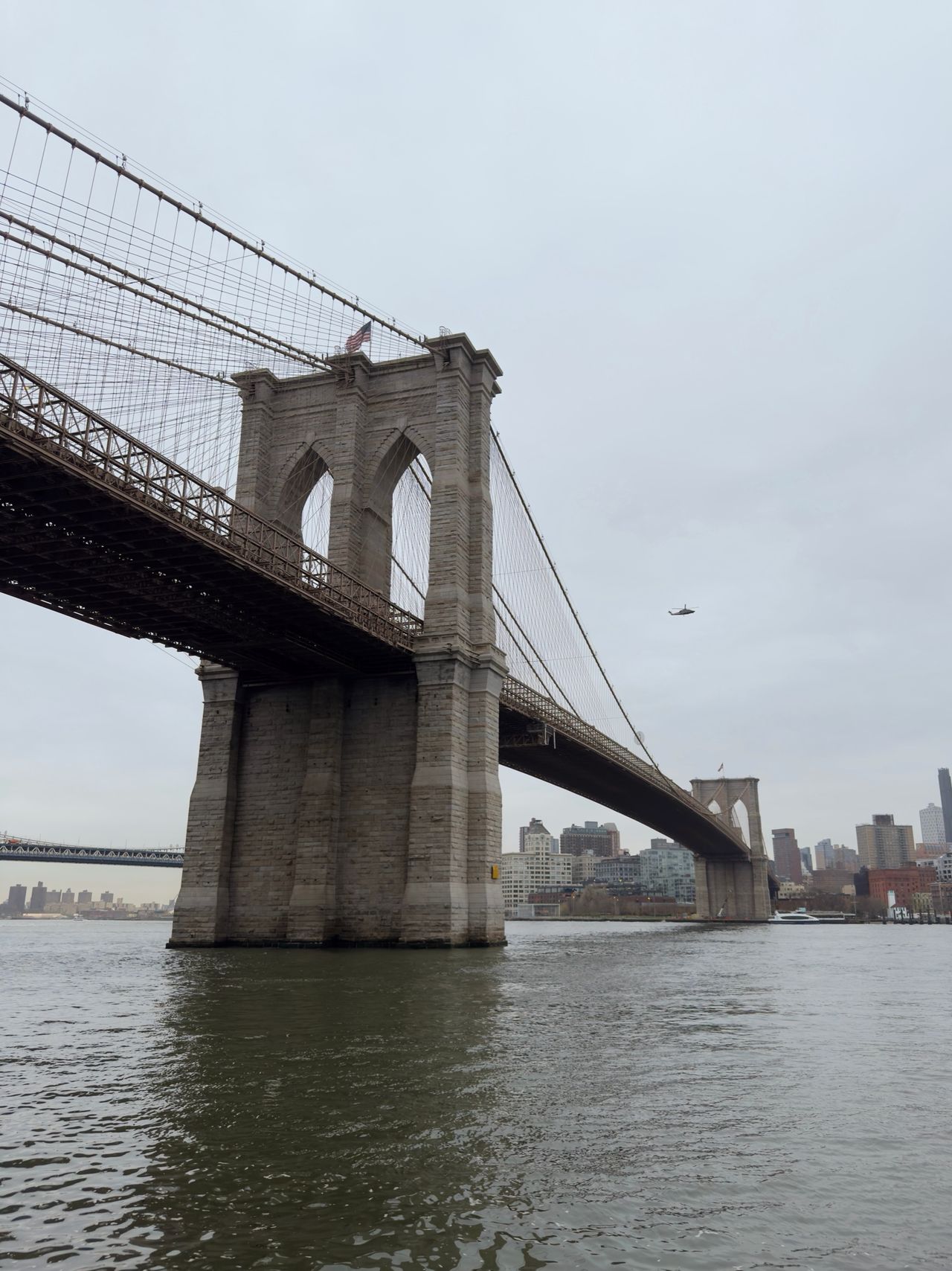 Brooklyn Bridge stone tower seen from below on the river, a helicopter hovering in the overcast sky beside it
