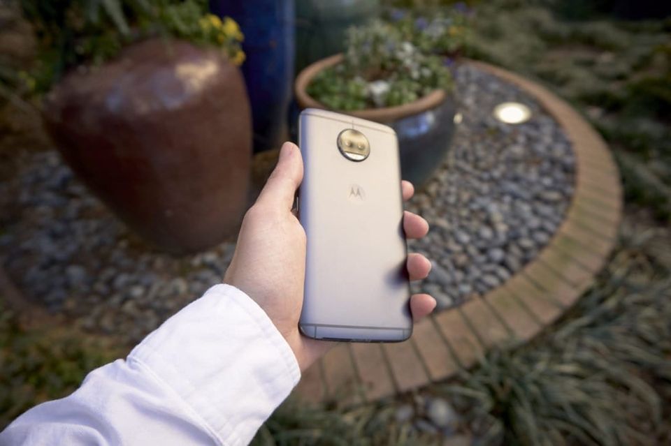 Hand holding a gold Motorola Moto G5s Plus phone, back facing up, over a garden with potted plants
