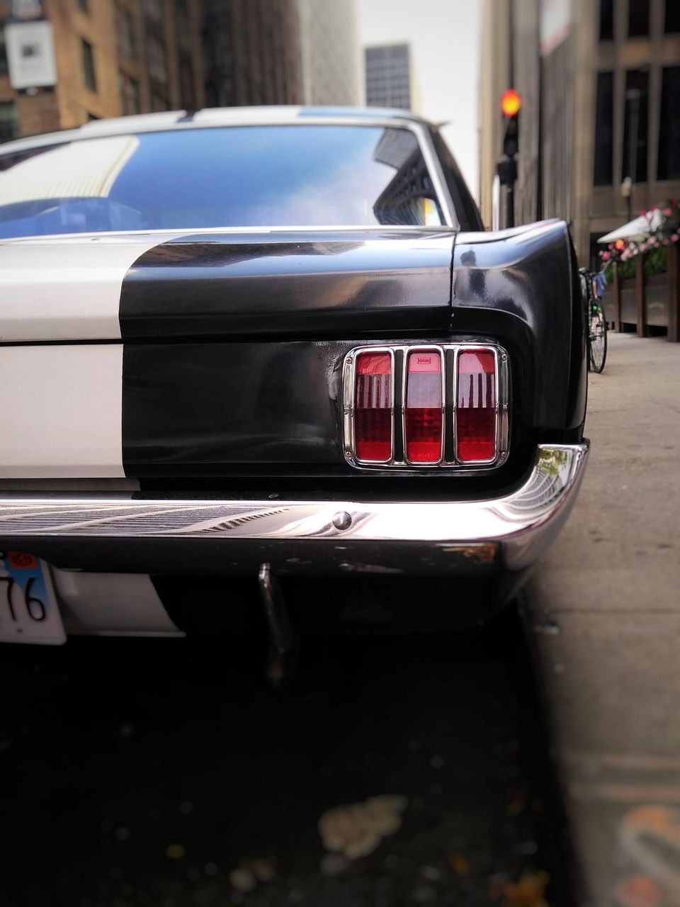 Rear taillight and chrome bumper of a black and white classic Mustang parked beside a city sidewalk