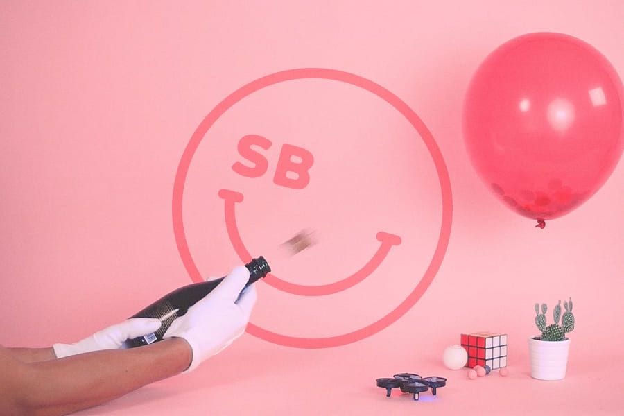 Gloved hands popping a champagne bottle in front of a pink SB smiley logo with a red balloon and a small drone