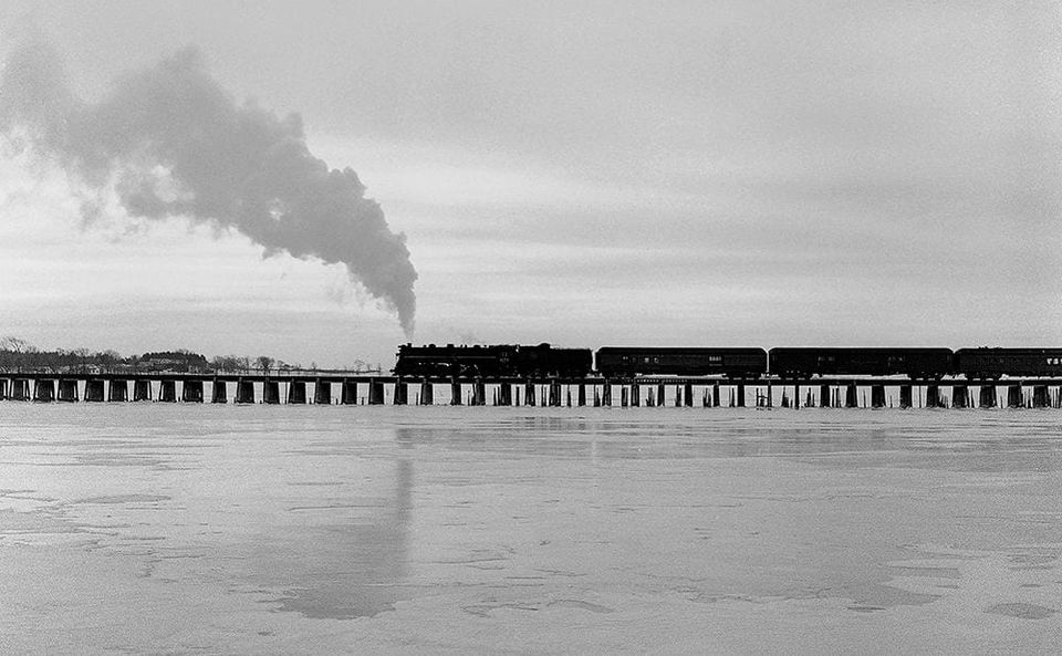 Black and white photo of a steam train crossing a long trestle over a frozen lake with smoke billowing