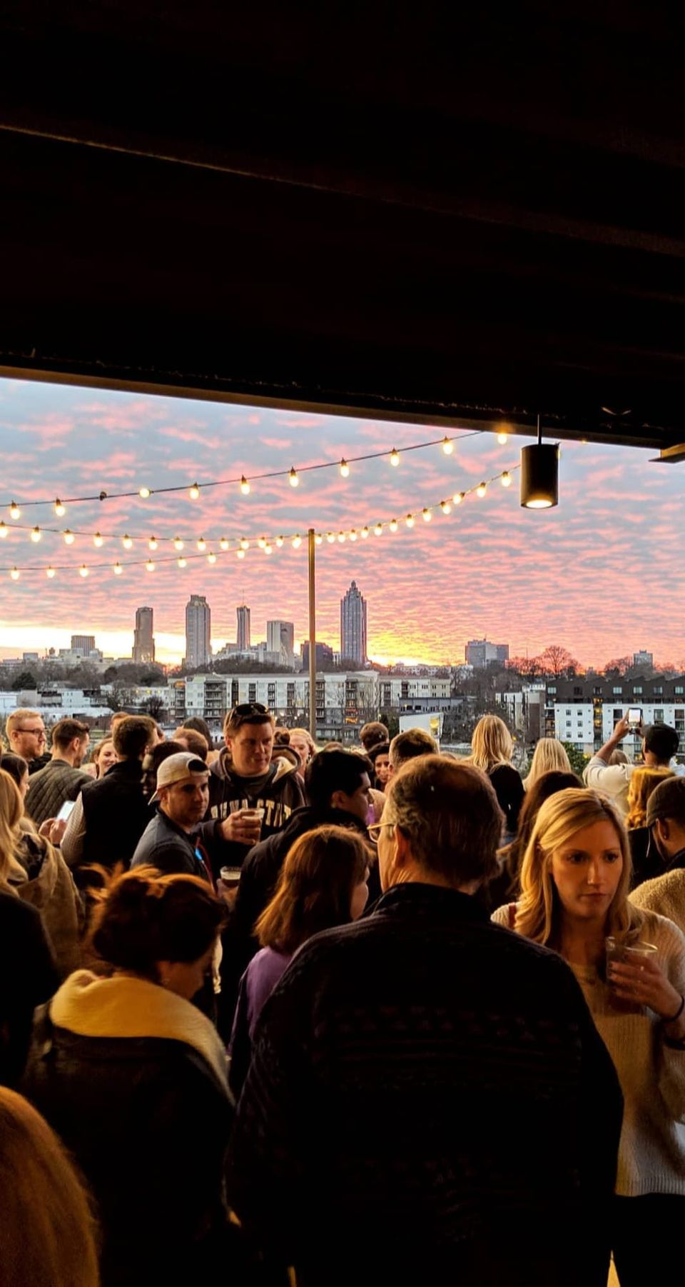 Crowd on a rooftop under string lights with a pink sunset sky over the Atlanta skyline