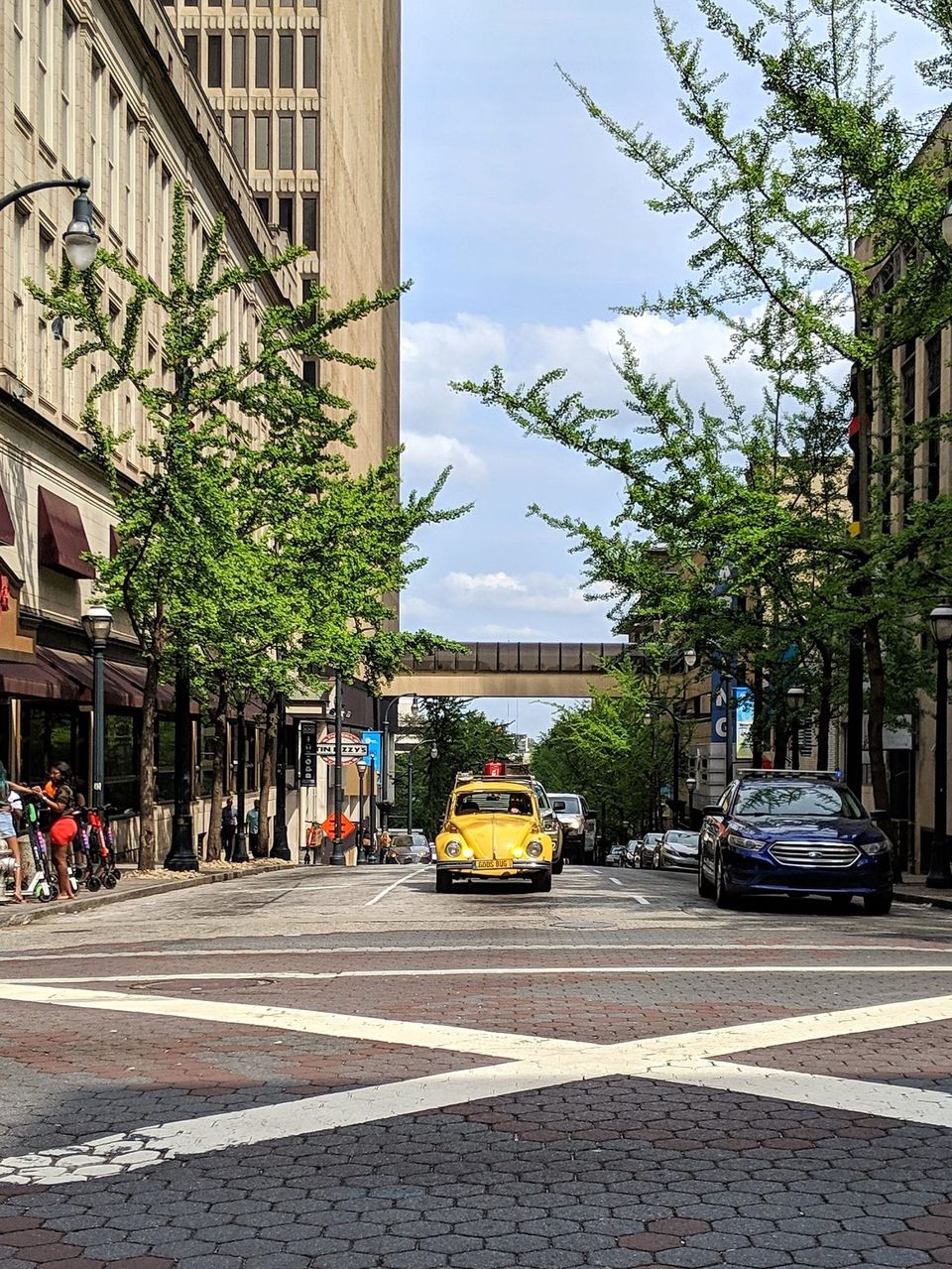 Yellow Volkswagen Beetle driving down a tree-lined downtown Atlanta street toward a skybridge