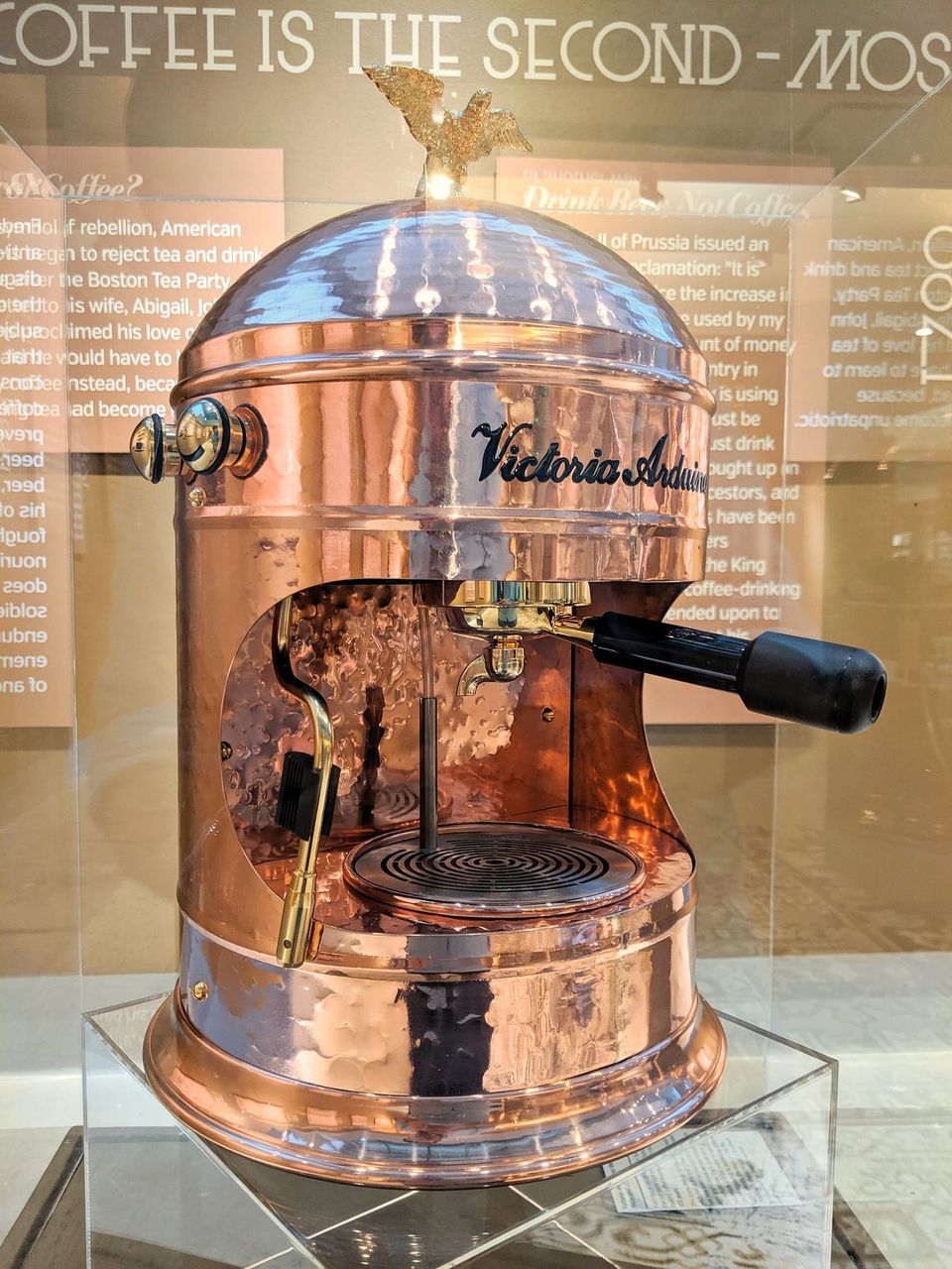 Hammered copper Victoria Arduino espresso machine topped with a gold eagle in a display case
