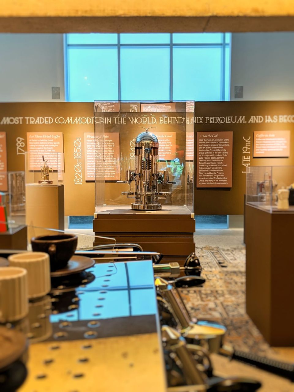 Tall chrome espresso machine in a glass case in a museum gallery of coffee history panels