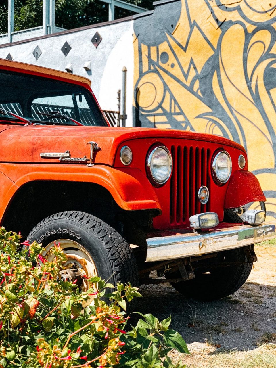 Front of a faded red Jeepster Commando parked beside a yellow and gray mural