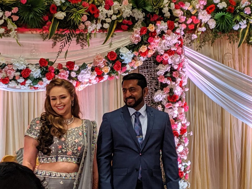 Smiling bride in a gray embellished lehenga beside a groom in a navy suit under a floral arch