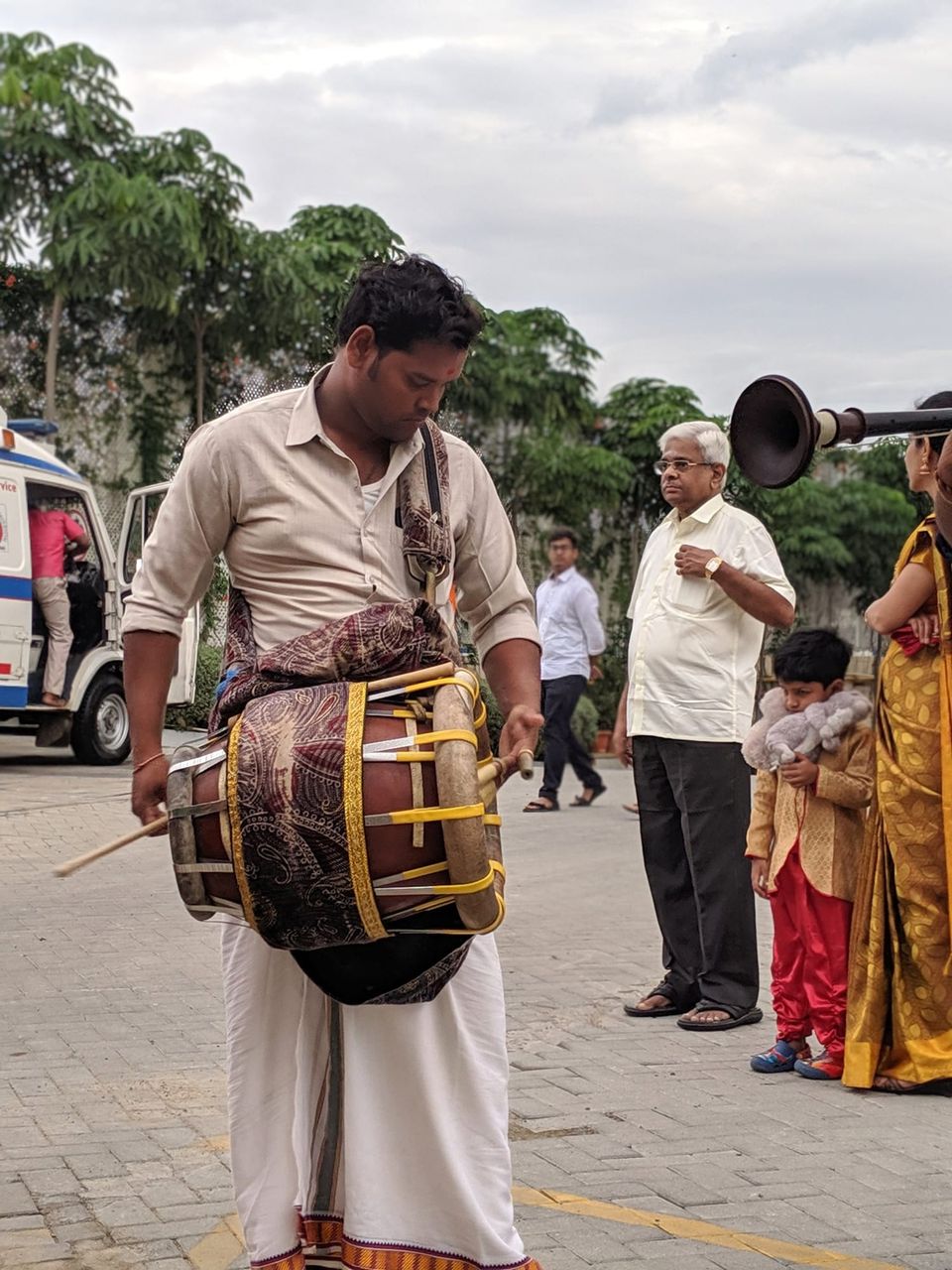 Drummer in a white dhoti playing a barrel drum as guests in traditional dress watch