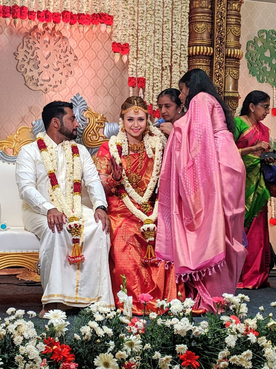 Bride in a red silk sari and jasmine garlands beside a groom in white on a decorated stage