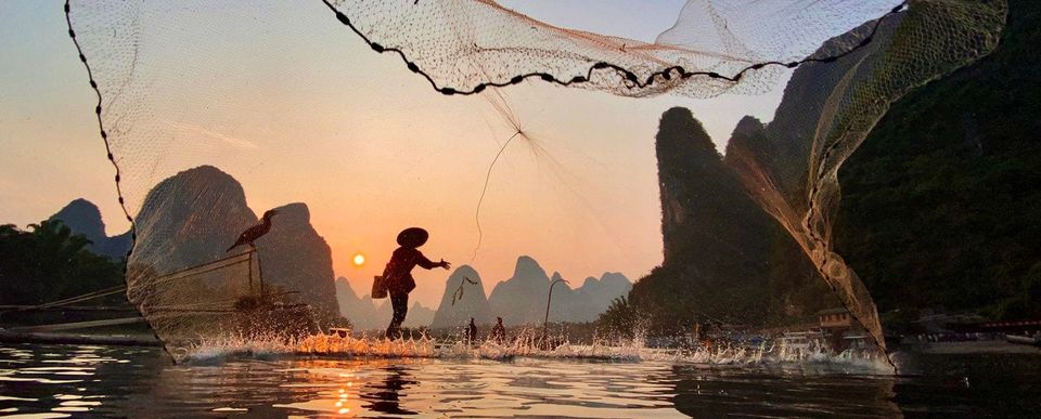 Silhouetted fisherman in a conical hat casting a net over a river at sunset among karst mountains