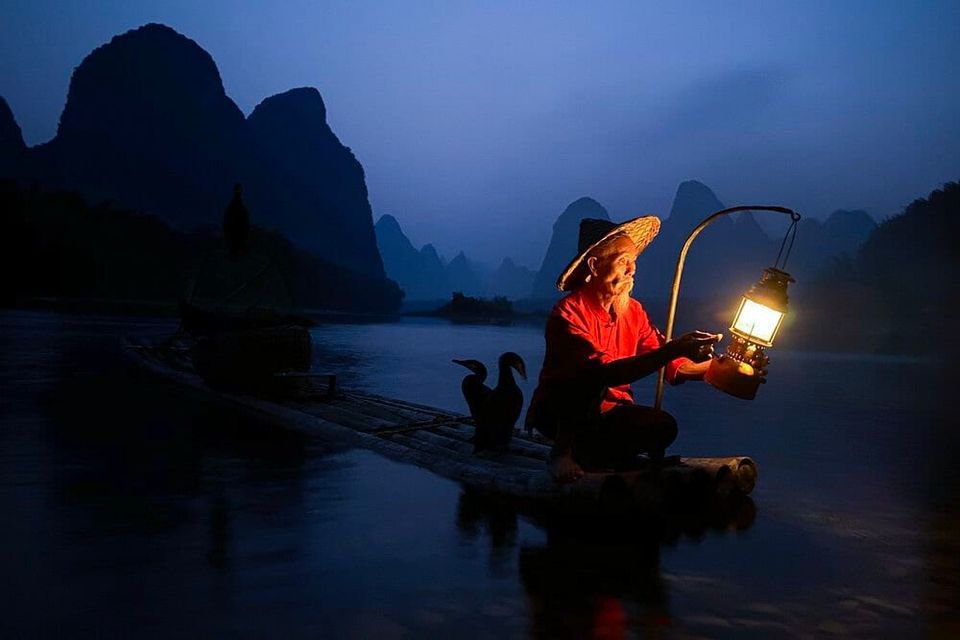 Elderly fisherman in a red shirt holding a glowing lantern on a bamboo raft with cormorants at dusk