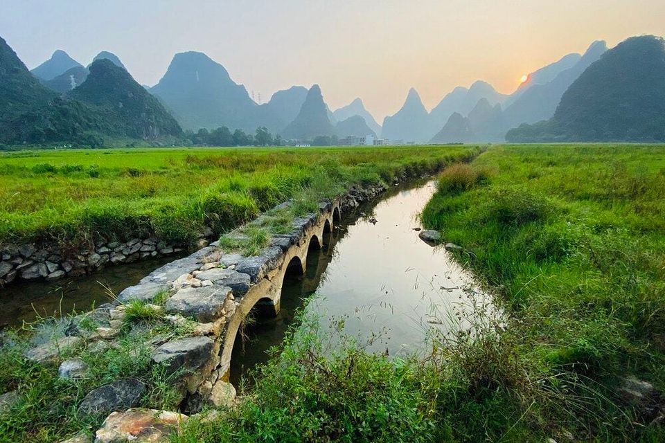 Stone arched bridge along a narrow canal through green fields toward karst peaks at sunset