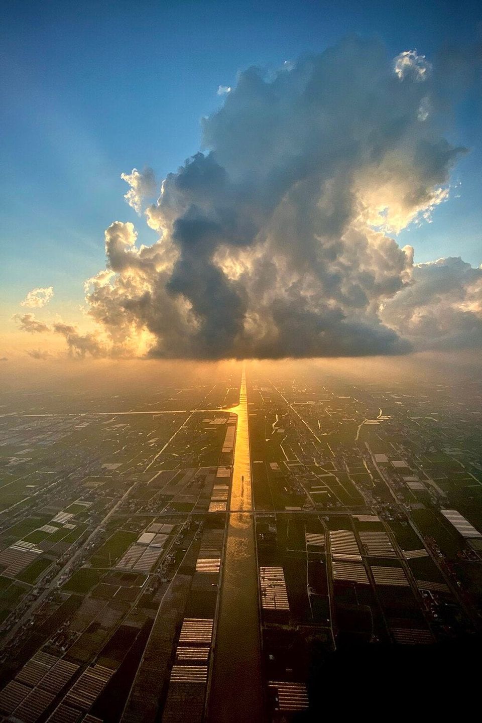 Aerial view of a sunlit canal stretching toward a towering cloud over farmland