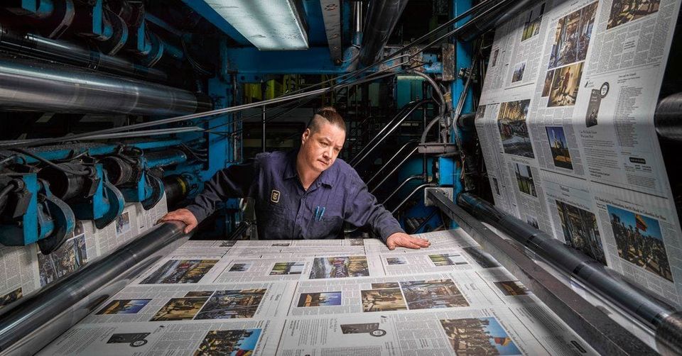 Pressman leaning over freshly printed newspapers running through a large blue printing press
