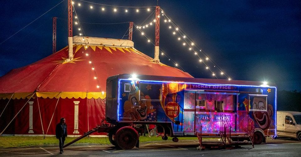 Red and yellow circus big top strung with lights at night beside a clown-painted ticket office trailer