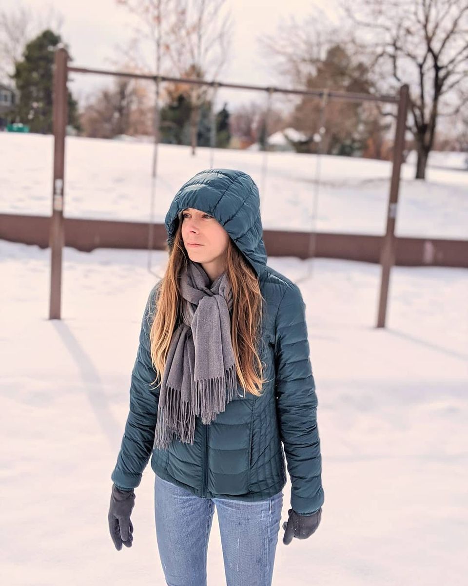 A woman with light-brown hair in a green hooded puffer jacket and gray scarf standing in a snowy park