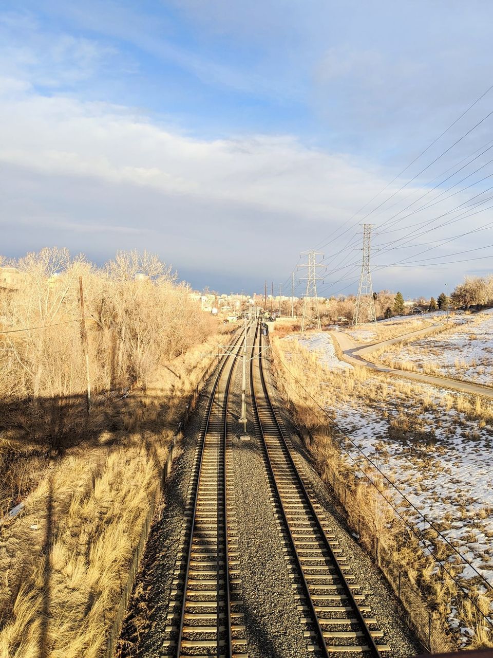 Twin light rail tracks curving into the distance past dry grass, snow and power line towers