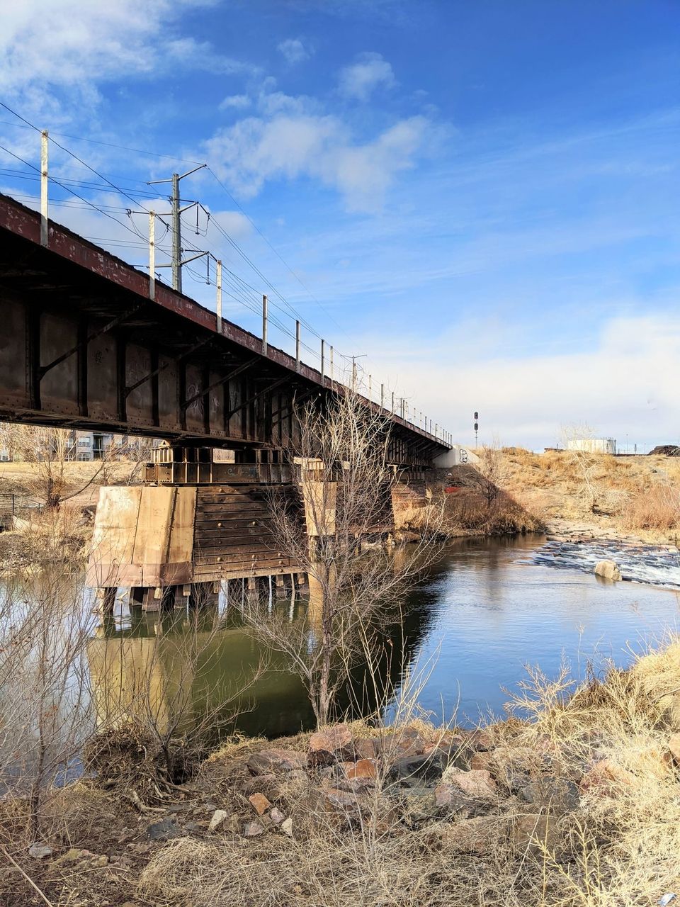 Rusty steel railroad bridge over a river with bare trees under a blue sky