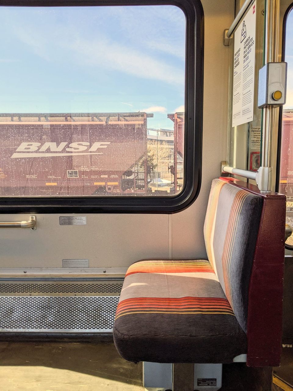 Striped light rail seat beside a window looking out on a maroon BNSF hopper car